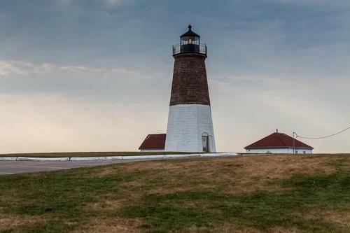 A tall lighthouse stands on a grassy hill, with two red-roofed buildings nearby under a cloudy sky.