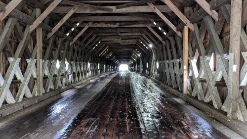 Interior of a covered bridge with wooden beams and a wet wooden floor, leading to bright light at the end.