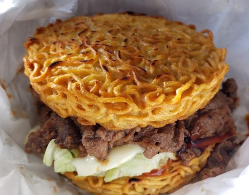 A burger with ramen noodle buns, filled with beef, lettuce, and sauces, wrapped in paper.