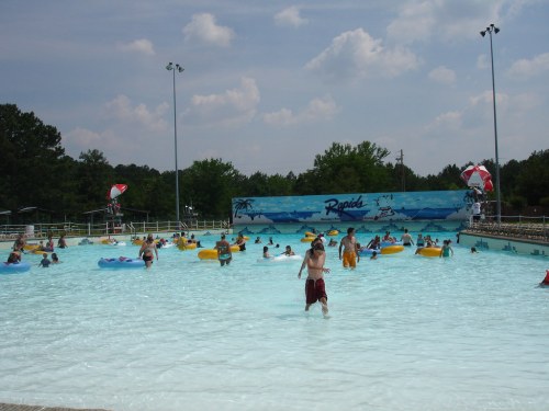 A busy water park wave pool with people swimming and floating on colorful rafts under a blue sky.
