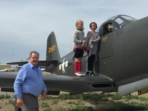 Two girls stand on a vintage military aircraft while a man smiles in front of them, with a cloudy sky in the background.