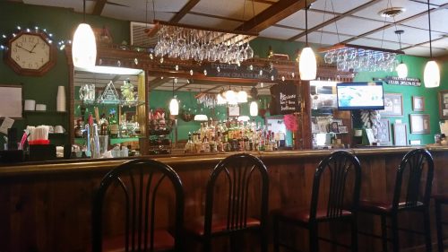 A cozy bar with a wooden counter, shelves of drinks, and hanging glasses, illuminated by warm lighting.