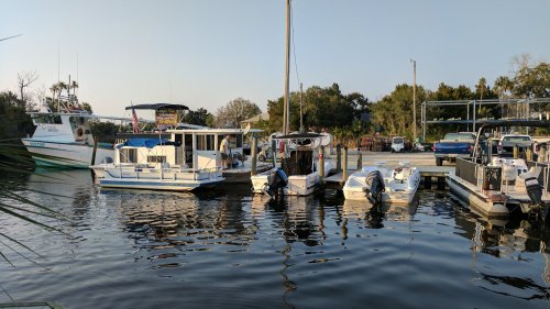 A marina scene with several boats docked in calm water, surrounded by trees and a parking area in the background.