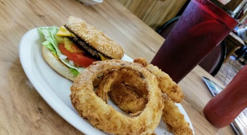 A plate with a sesame seed burger topped with lettuce, tomato, and cheese, alongside crispy onion rings and a drink.