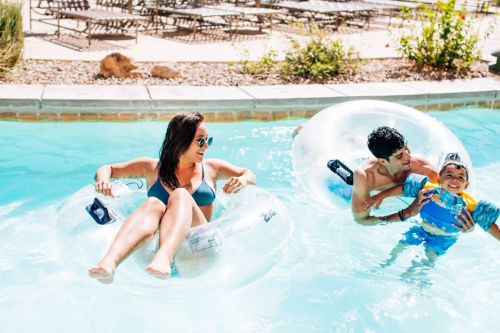 A woman relaxes on a float in a pool while two boys play nearby, enjoying a sunny day.