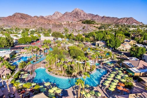 Aerial view of a resort with pools, palm trees, and mountains in the background under a clear blue sky.