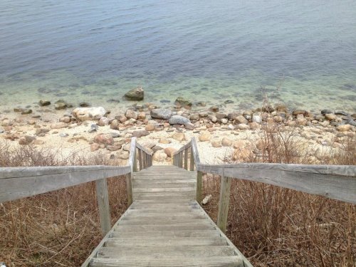 Wooden steps lead down to a rocky shoreline with clear water and pebbles along the beach.