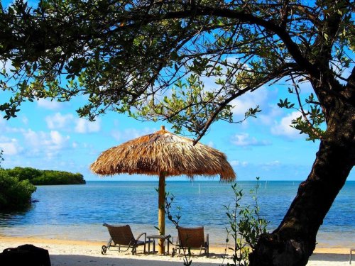 A serene beach scene with a thatched umbrella, two chairs, and calm waters under a blue sky with scattered clouds.