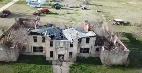 Aerial view of a burned-out, abandoned building surrounded by overgrown grass and vehicles in the background.