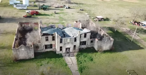 Aerial view of a dilapidated, burned-out building surrounded by open fields and scattered vehicles.