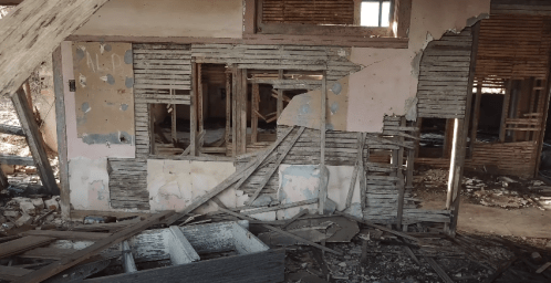 An abandoned room with crumbling walls, broken windows, and scattered debris on the floor.