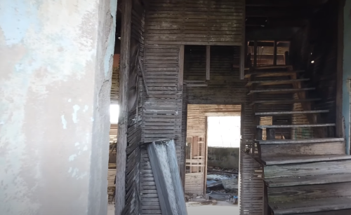 Interior of an abandoned building, featuring wooden walls, a staircase, and debris scattered on the floor.