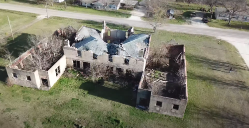 Aerial view of a dilapidated, abandoned building with a collapsed roof, surrounded by overgrown grass and nearby road.