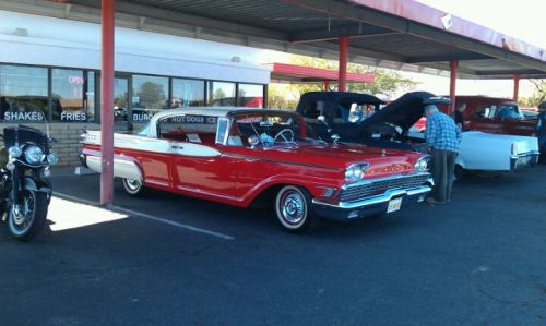 A classic red car parked outside a diner, with people and other vintage vehicles nearby.