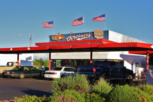 A retro diner with American flags, cars parked outside, and a large sign reading "Screamer's" against a clear blue sky.