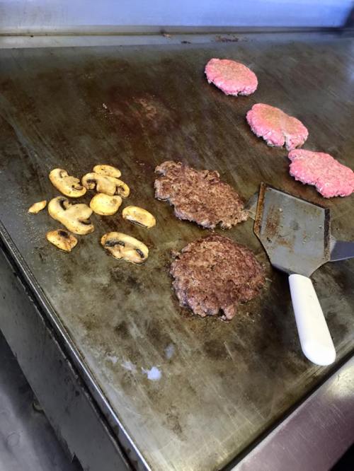 Griddle with cooking hamburger patties and sliced mushrooms, with a spatula resting nearby.