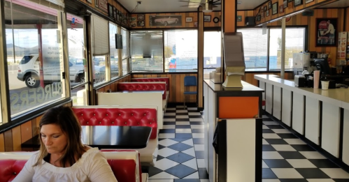 A retro diner interior with red booths, checkered floor, and a woman sitting at a table, looking down.