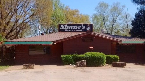Exterior view of Shane's Restaurant, featuring a brick facade and green awning, surrounded by trees and shrubs.