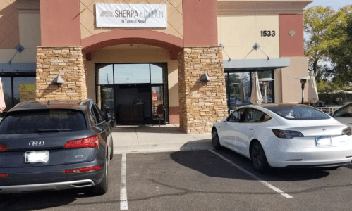 Exterior of Sherpa Kitchen restaurant with a sign, two parked cars in front, and a clear blue sky.