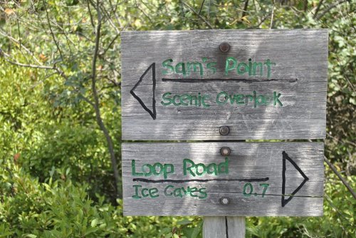 Wooden signpost with directions to Sam's Point, Scenic Overlook, Loop Road, and Ice Caves, surrounded by greenery.