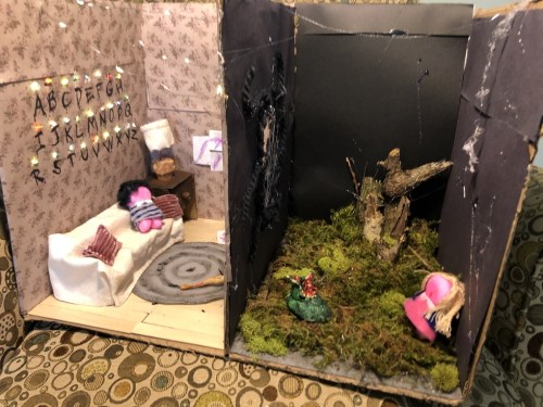 A diorama featuring two rooms: one with a sofa and letters on the wall, the other with moss and a small tree.