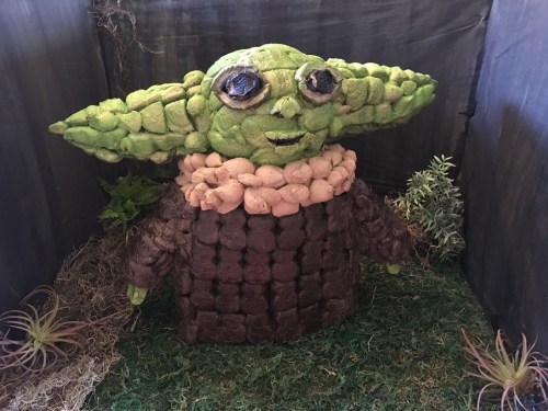 A whimsical sculpture of a green character with large ears, made from textured materials, set in a natural-themed display.