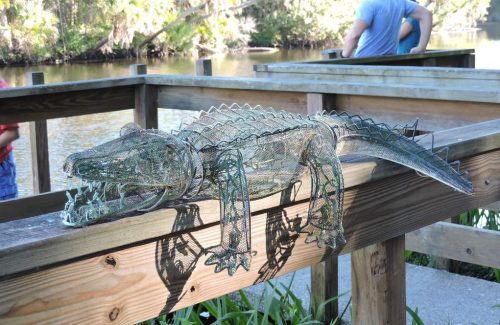 A wire sculpture of an alligator resting on a wooden railing by a riverbank, surrounded by greenery.