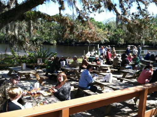 A busy outdoor dining area by a river, with people sitting at picnic tables enjoying food and conversation.