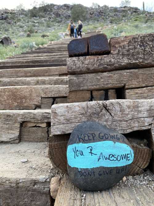 A painted rock on wooden steps reads, "Keep going! You’re awesome! Don’t give up!" with hikers in the background.