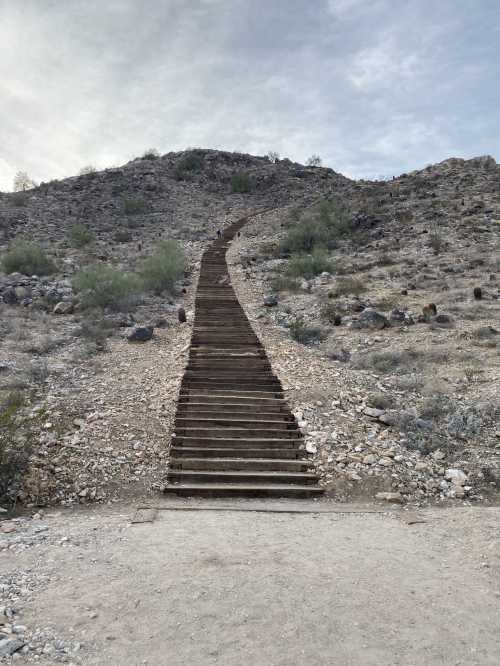 A long, winding staircase leads up a rocky hillside under a cloudy sky. Sparse vegetation surrounds the path.