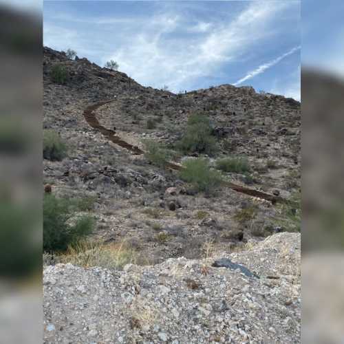 A winding dirt path leads up a rocky hillside under a blue sky with scattered clouds.