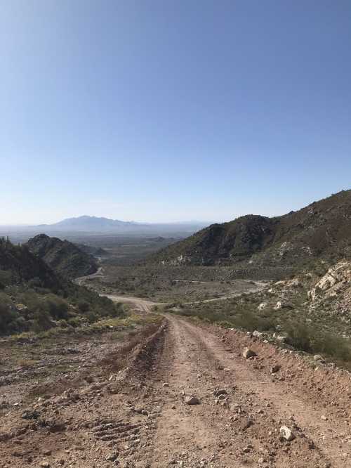 A dirt road winds down a mountain, with a vast valley and distant mountains visible under a clear blue sky.