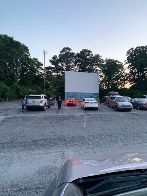 A drive-in movie theater with parked cars facing a large blank screen, surrounded by trees at dusk.