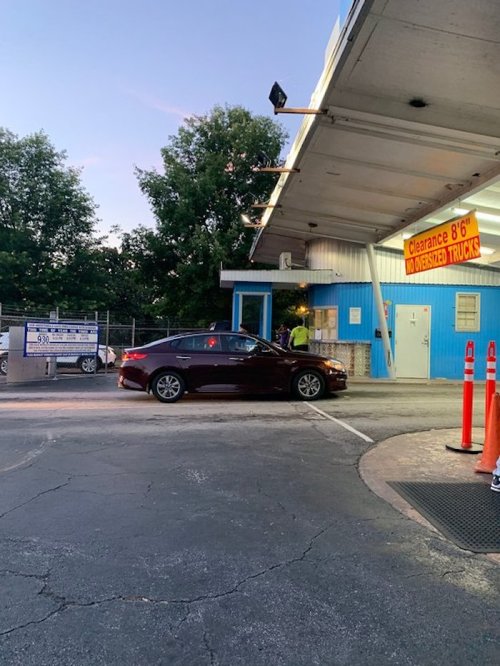A car approaches a toll booth with a clearance sign for oversized trucks, surrounded by trees and orange cones.