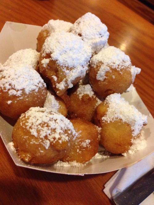 A serving of golden-brown, powdered sugar-dusted doughnut holes in a white paper tray.