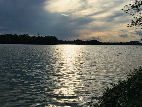 A serene lake at sunset, reflecting soft light and clouds, with trees lining the shore.