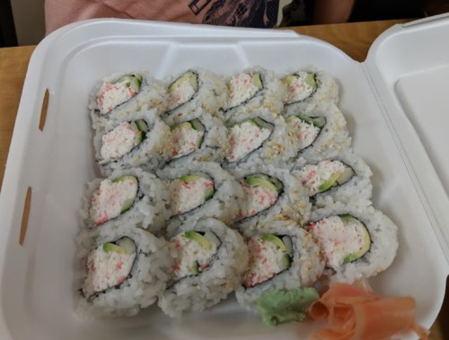 A takeout container filled with neatly arranged sushi rolls, featuring crab, cucumber, and wasabi on the side.