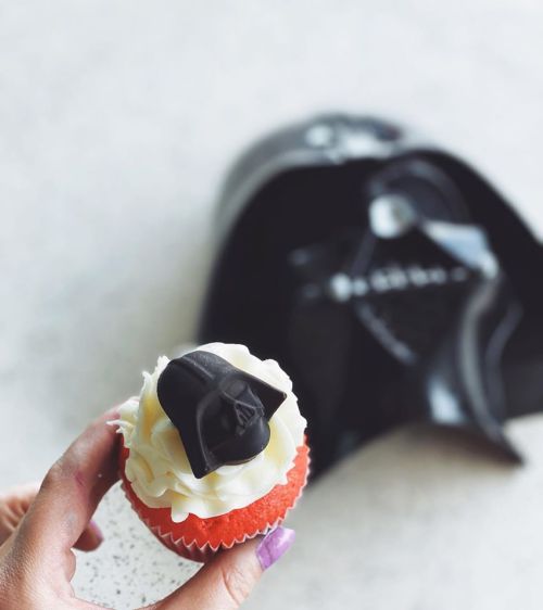 A hand holding a red cupcake topped with white frosting and a black Darth Vader figure, with a Darth Vader bag in the background.