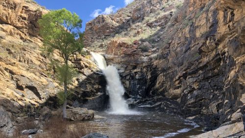 A waterfall cascades down rocky cliffs into a serene pool, surrounded by rugged terrain and sparse vegetation.