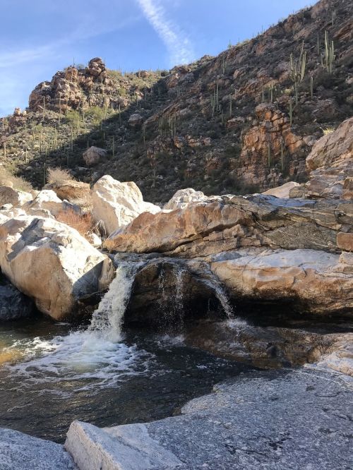 A serene waterfall cascades over rocks into a clear pool, surrounded by rugged mountains and cacti under a blue sky.