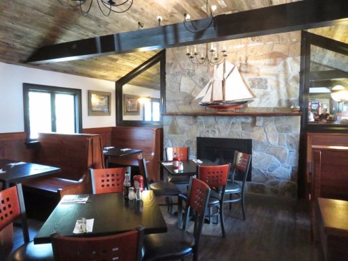 Cozy restaurant interior with wooden booths, a stone fireplace, and a ship model on the wall. Warm, inviting atmosphere.