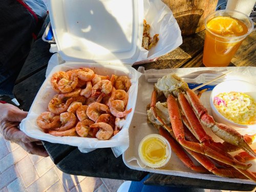 A takeout meal featuring shrimp, crab legs, coleslaw, and a drink on a wooden table.