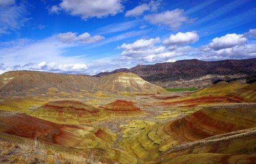 Colorful hills and valleys under a blue sky with clouds, showcasing vibrant red, yellow, and green layers of rock.