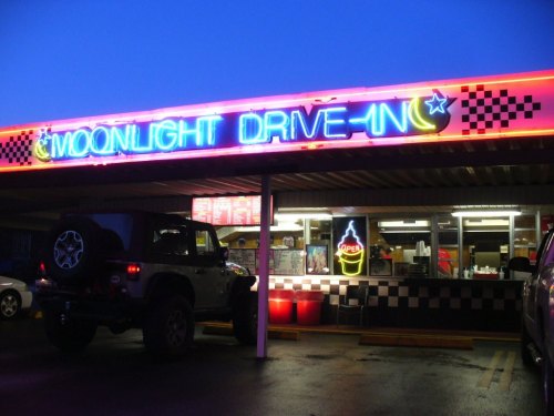 Neon sign of "Moonlight Drive-In" with a vintage car parked outside, illuminated at dusk.