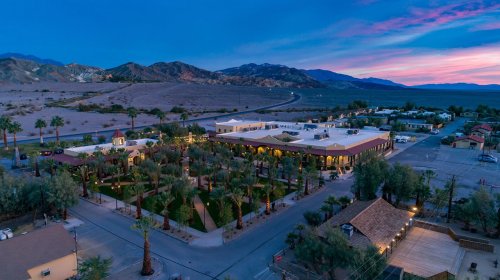 Aerial view of a desert resort with palm trees, mountains in the background, and a colorful sunset sky.