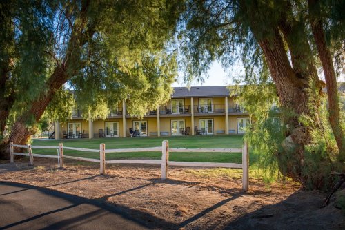A yellow building with balconies surrounded by trees and a grassy area, viewed through a wooden fence.