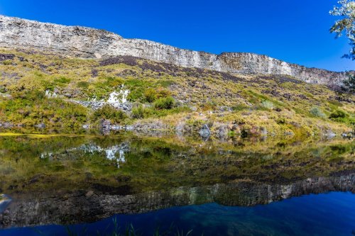A serene landscape featuring a rocky cliff reflected in calm water, surrounded by lush greenery under a clear blue sky.