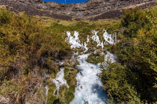 A cascading waterfall flows through lush greenery, surrounded by rocky cliffs under a clear blue sky.