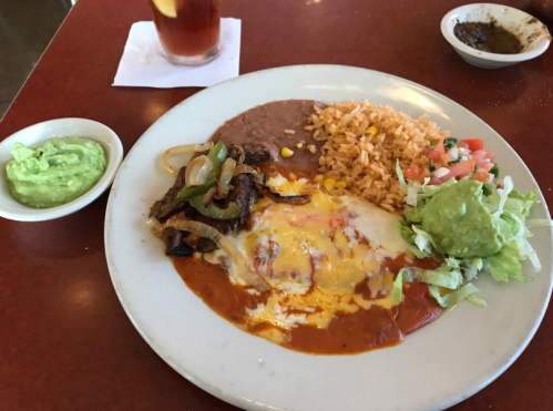 A plate of enchiladas topped with cheese, served with rice, refried beans, salad, and guacamole on the side.
