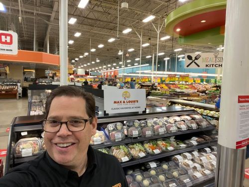 A smiling man stands in a grocery store aisle, showcasing prepared meals and fresh produce in the background.